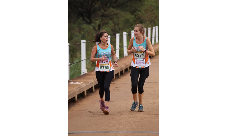 Two women in athletic wear are running together on a path, chatting and smiling. They wear race bibs, suggesting a marathon or race event. Trees line the background.