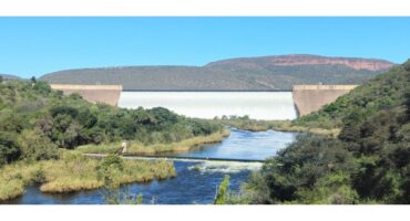A wide dam with cascading water sits between two hills, surrounded by lush greenery. A river flows in the foreground under a clear blue sky.
