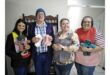 Four smiling people stand together holding knitted teddy bears and clothing. The group appears cheerful and proud of their handmade crafts.