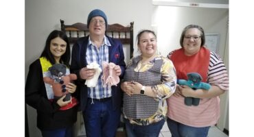 Four smiling people stand together holding knitted teddy bears and clothing. The group appears cheerful and proud of their handmade crafts.