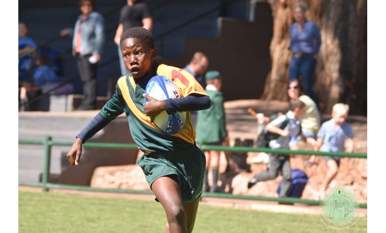A young rugby player in yellow and green sprints with determination, holding a rugby ball. The background shows spectators and trees under bright sunlight.