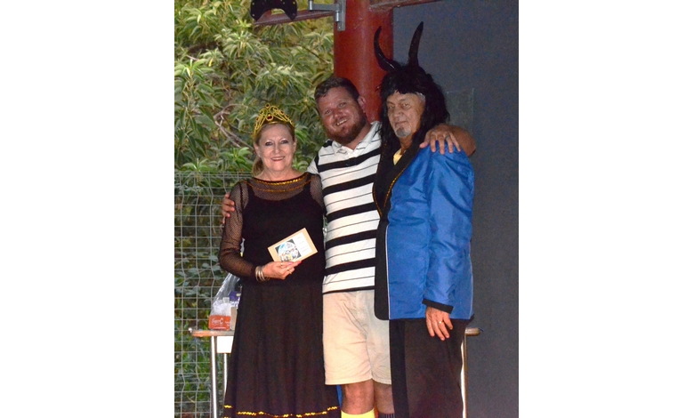 Three people in costume pose together. The left person wears a black dress and crown, holding a card. The middle person smiles in a striped shirt. The right person, dressed in a blue jacket and horned headpiece, has an arm around the middle person. They stand in front of a metal fence with greenery behind it.