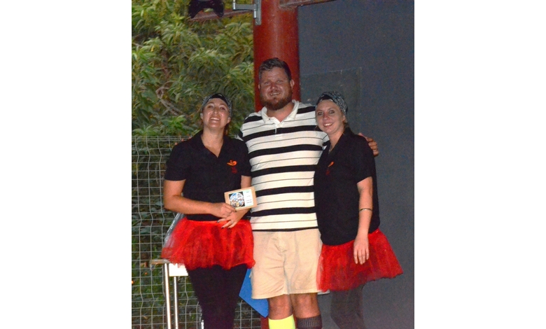 Three people pose together outdoors, smiling. Two wear black shirts and red tutus, while the man wears a striped shirt and shorts. The mood is cheerful.