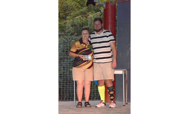 Two people stand smiling under a tree. The person on the left holds a small item. Both wear casual shorts; one has vibrant mismatched socks, creating a fun, relaxed atmosphere.