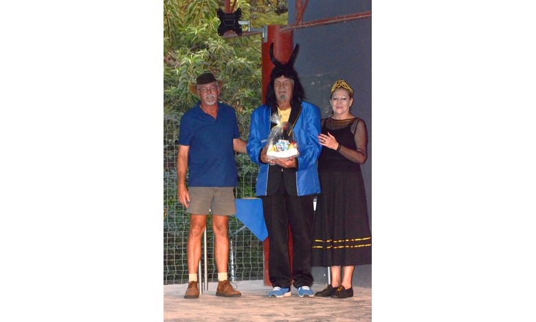 Three people on a stage, one in a blue jacket with black horns holding a gift basket, flanked by a man in casual attire and a woman in a black dress.