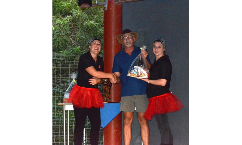 Two women in black tops and red skirts stand beside a man in casual clothes and a hat, smiling and holding a gift basket, outdoors with greenery.