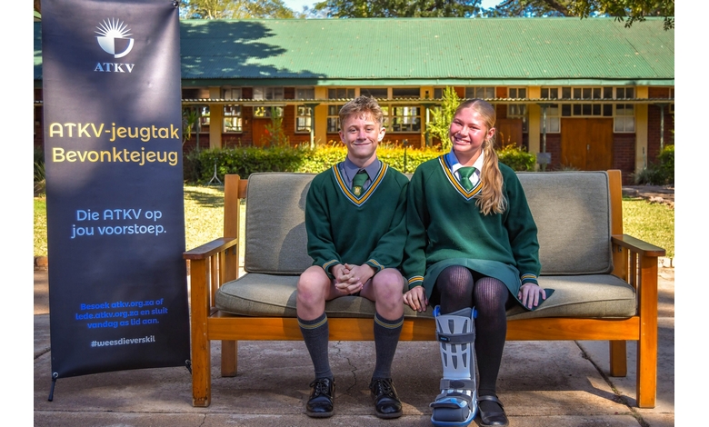 Two young students seated on a bench, with a visible sign that reads "ATKV," looking cheerful and relaxed.