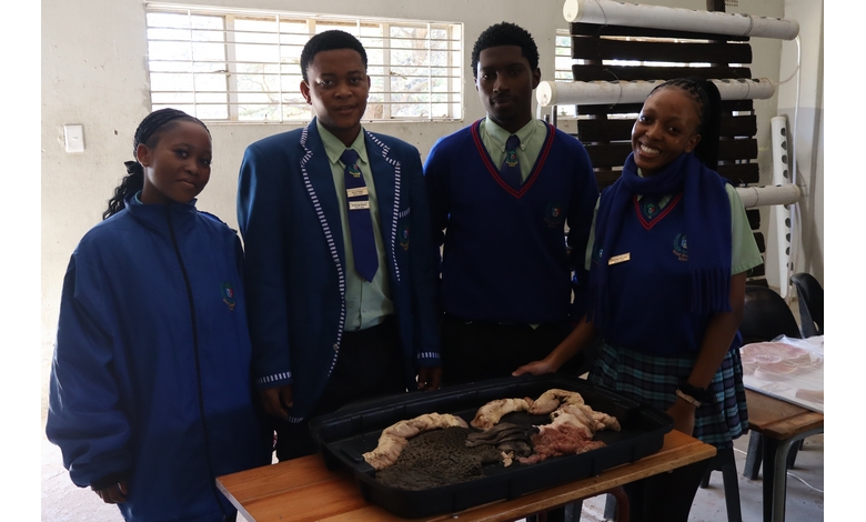 Four students in blue school uniforms stand around a table with soil and organic material. They appear proud and collaborative in a classroom setting.