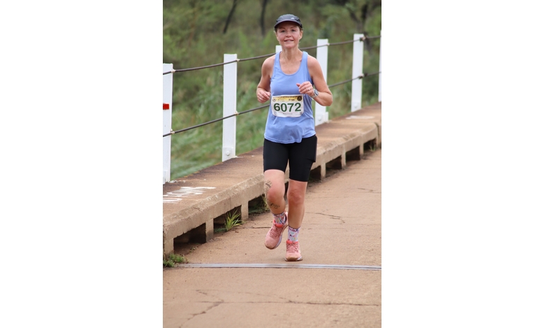 A woman in a blue tank top and black shorts runs outdoors on a paved path. She wears a race bib numbered 6072, a black cap, and pink shoes, looking focused.