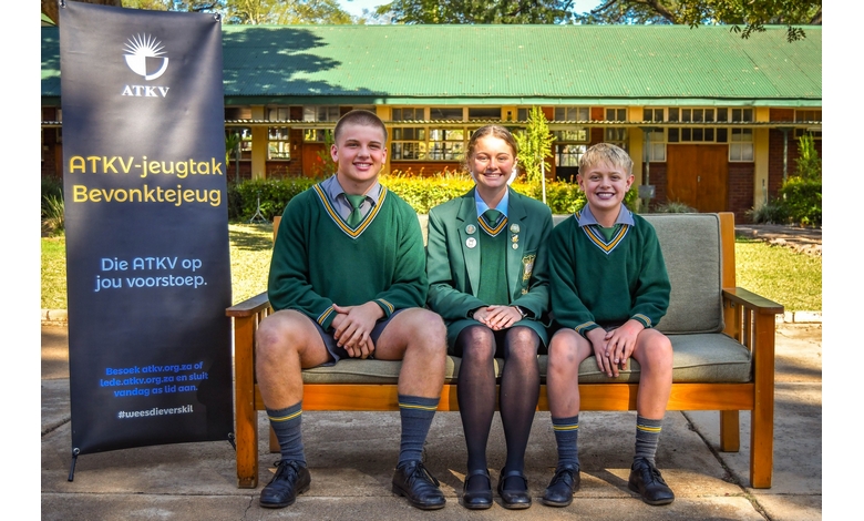 Three students are seated on a bench, with a visible sign that says "ATKV" in the background.