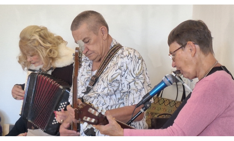Three elderly musicians perform together. One plays an accordion, another a guitar, and the third sings into a microphone. The scene is warm and engaging.