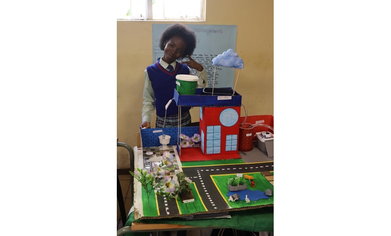 A student proudly presents a colorful eco-friendly city model with greenery, roads, a water body, and a rainwater harvesting system. The mood is creative and educational.