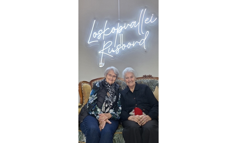 Two elderly women sit close together on a vintage sofa, smiling warmly. A neon sign above them reads "Loskopvallei Rusoord." The atmosphere is cozy and joyful.