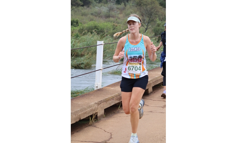 Runner in a turquoise tank top and navy shorts jogs on a scenic riverside path. She wears a visor, displaying focus and determination.