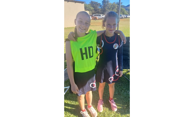 Two young girls wearing netball dresses stand on a grassy field, smiling and with arms around each other. One wears a bright bib marked "HD".