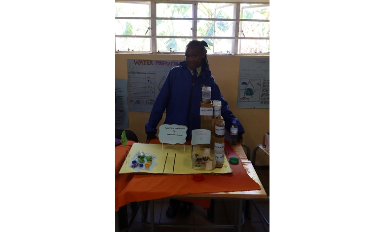 A student in a blue uniform stands behind a display table with water purification bottles and colorful samples. Posters hang on the beige wall behind.