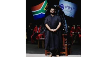 A man wearing black graduation robes stands on stage, facing the audience with a serious expression.