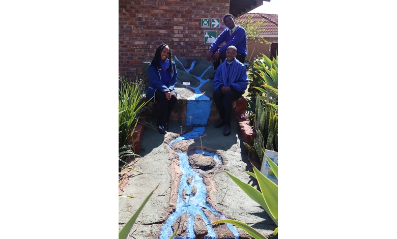 Three people wearing blue uniforms sit and stand on a model of a hill with a painted blue river. They are smiling, surrounded by green plants.