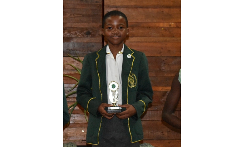 A student in a dark green blazer with yellow trim proudly holds a trophy. The background is wooden. The mood is celebratory and proud.