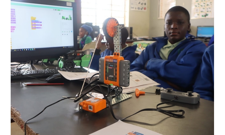 A student in a blue jacket sits at a desk with a robotics kit, a computer displaying a coding program, and various electronics, focused and engaged.