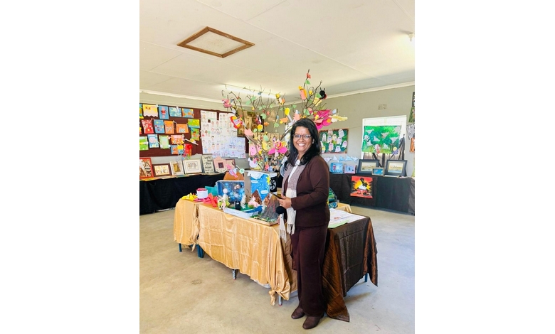 A person stands smiling in an art exhibit room. They hold a mug next to a table with colorful decorations and children's artwork. The atmosphere is lively and creative.