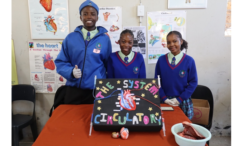 Three students in blue uniforms stand behind a science project display titled "The Circulatory System." They appear enthusiastic and proud.