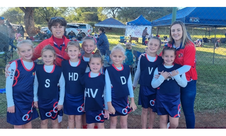 A girls' sports team poses with two coaches outdoors. The players wear navy uniforms with initials, smiling under sunny skies and blue tents in the background.