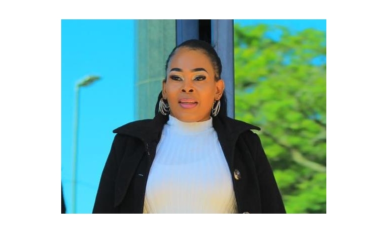A woman in a white turtleneck and black coat stands outside. She wears large hoop earrings. The background features green foliage and a clear blue sky.