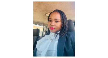 A woman with straight black hair and red lipstick wears a white ruffled blouse and black jacket, sitting confidently in a car, conveying professionalism.