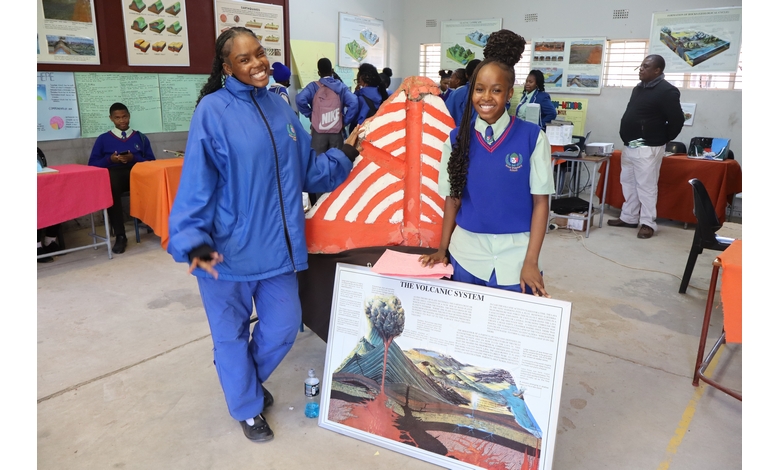 Two students in a classroom smile beside a volcano model and a poster titled "The Volcanic System." The setting is lively with several students in the background.