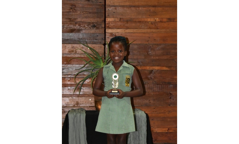 A smiling young girl in a green checkered school uniform holds a trophy in front of a wooden wall, conveying pride and achievement.