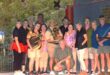 A group of thirteen people, dressed in casual and colorful attire, pose together in front of lush greenery. One person kneels in front, holding a trophy. The mood is joyful and celebratory, suggesting a team event or competition.