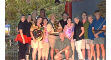 A group of thirteen people, dressed in casual and colorful attire, pose together in front of lush greenery. One person kneels in front, holding a trophy. The mood is joyful and celebratory, suggesting a team event or competition.