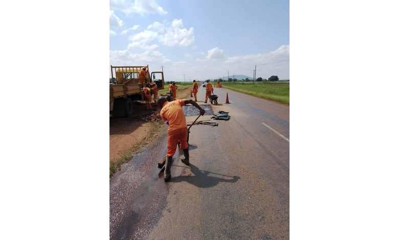 Workers in orange uniforms repair a rural road under a partly cloudy sky. Some use tools while others rest near a parked truck and orange cones.