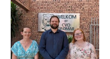Three people stand smiling in front of a brick wall with a sign saying “Welkom by CVO Gromar.” The atmosphere is friendly and welcoming.