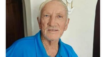 Elderly man with short white hair and a light beard wearing a bright blue polo shirt, smiling gently; relaxed, warm atmosphere.