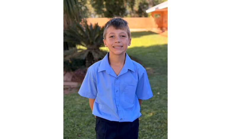 A young boy with short hair smiles, wearing a light blue short-sleeve shirt, standing on green grass with a blurred garden and trees in the background.