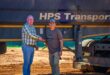 Two men shake hands in front of a large truck with "HPS Transport" on it. One wears a plaid shirt, the other a cap and t-shirt. The setting is sunny and sandy.