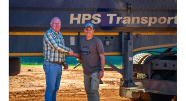 Two men shake hands in front of a large truck with "HPS Transport" on it. One wears a plaid shirt, the other a cap and t-shirt. The setting is sunny and sandy.
