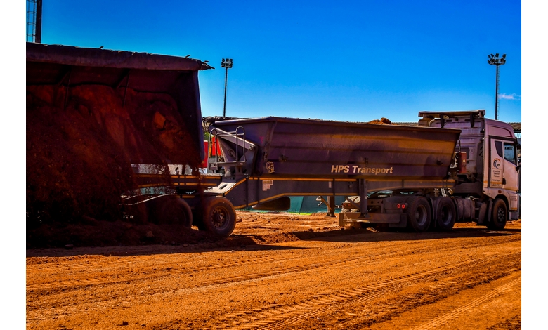 A large truck with "HPS Transport" on the side hauls dirt on a sunny construction site with a vivid blue sky and towering floodlights.