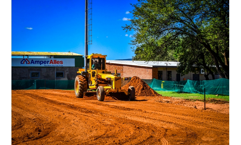 A yellow bulldozer shapes red soil under a vibrant blue sky. Nearby, a building with a logo reads "Amper Alles," and green trees provide shade.
