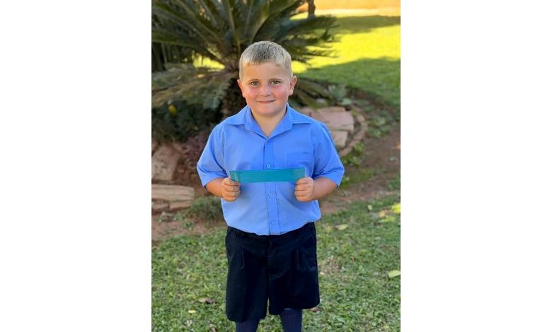 A young boy in a blue shirt and dark shorts smiles while holding a colorful strip outdoors. He stands on green grass with plants in the background.