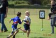 Two young boys play rugby on a grassy field. One holds the ball while the other chases him. A parent holds a child nearby. The scene is lively and energetic.