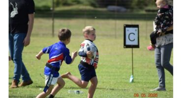 Two young boys play rugby on a grassy field. One holds the ball while the other chases him. A parent holds a child nearby. The scene is lively and energetic.
