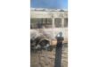 Charred remains of a bus with windows missing and ash covering the exterior. A person's shadow is cast on the bus, suggesting a scene of destruction.