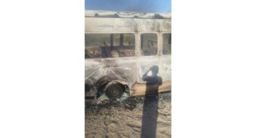 Charred remains of a bus with windows missing and ash covering the exterior. A person's shadow is cast on the bus, suggesting a scene of destruction.