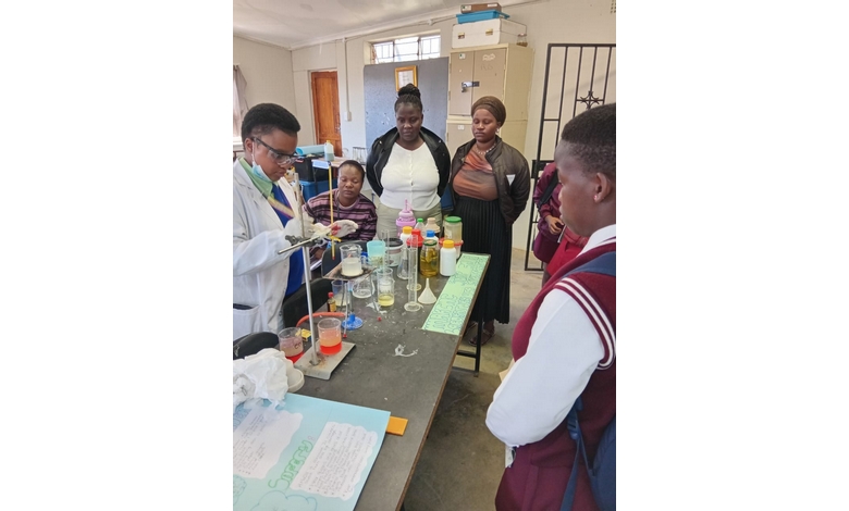 A science teacher in a lab coat conducts an experiment for attentive students in a classroom. The table is filled with beakers of colorful liquids. The atmosphere is educational and focused.
