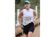 Woman in a white tank top and visor runs outdoors, wearing a race bib numbered 6353. She appears focused and determined, with a blurred green background.