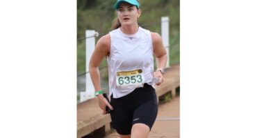 Woman in a white tank top and visor runs outdoors, wearing a race bib numbered 6353. She appears focused and determined, with a blurred green background.