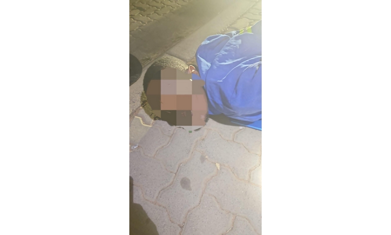 A man wearing a blue shirt lying face down on the pavement. The visible part of his face is blurred out.